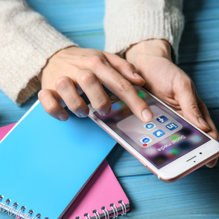 Main tenant un téléphone pour cliquer sur les réseaux sociaux. Fond bleu, carnets posés sur le bureau. Algorithme linkedin